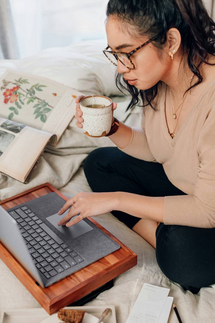 young, dark haired woman wearing glasses, working on laptop while drinking coffee
